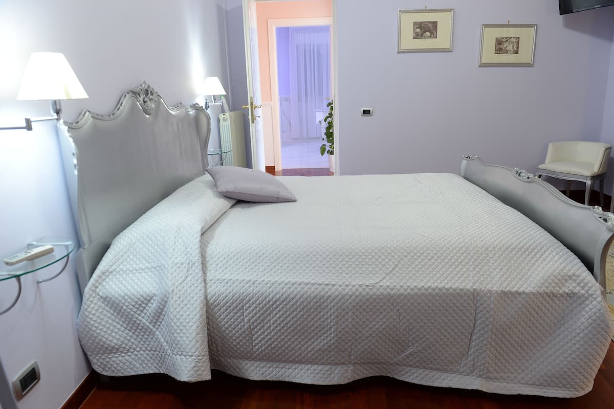Una cama bien hecha con cubrecama blanco acolchado, una mesita de noche con lámpara, y una silla a la derecha.