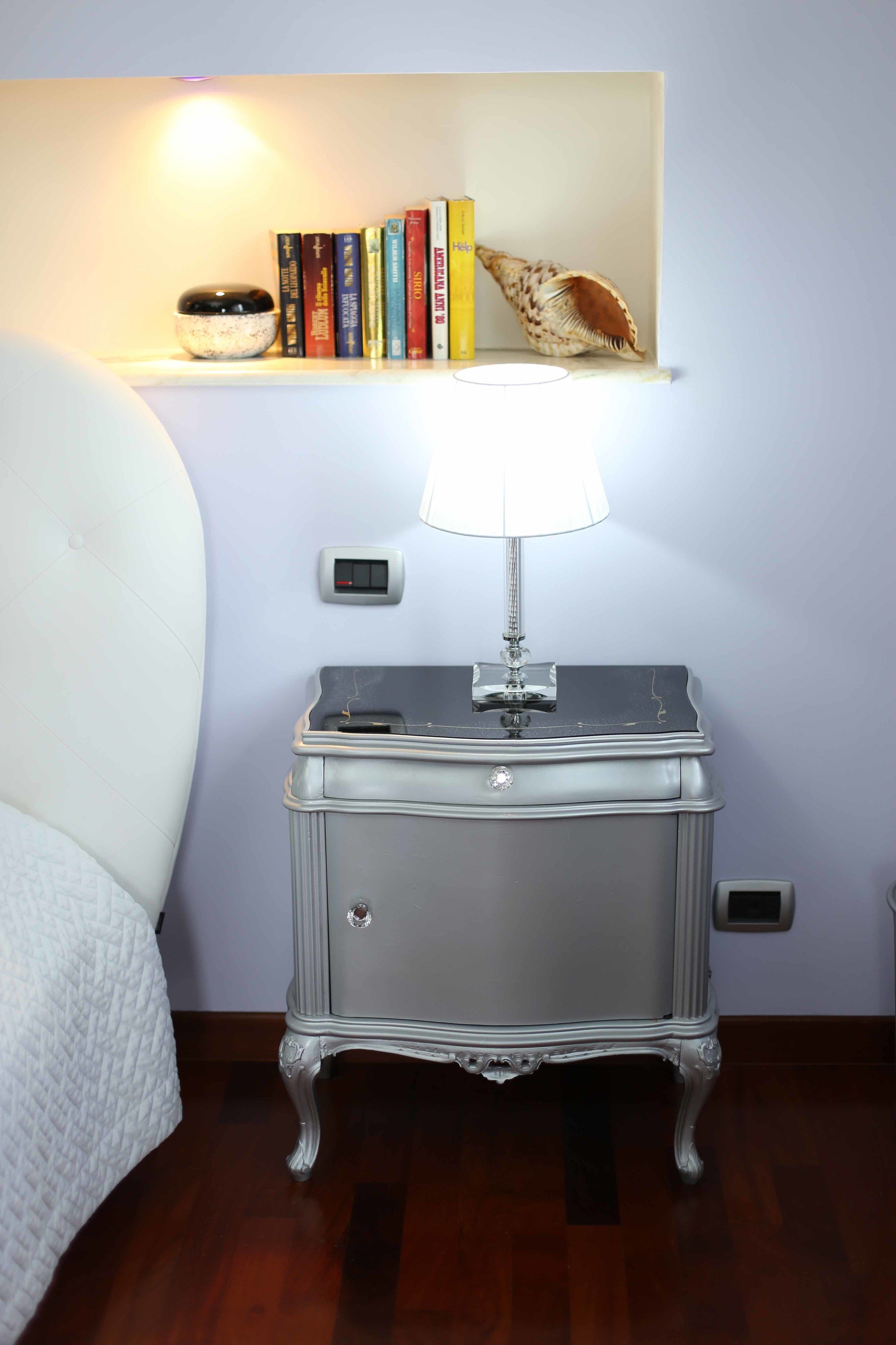 A bedroom with a white bed, a nightstand with a lamp, a shelf with books and a seashell, and a wall-mounted light.