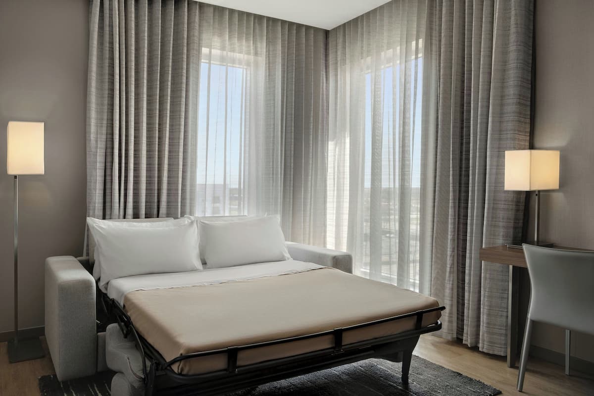 Ein modernes Hotelzimmer mit Bett, Nachttischen und Blick auf die Stadt durch transparente Vorhänge.