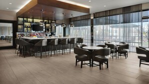 Zona de bar moderna con mostrador, varios taburetes y mesas, con grandes ventanales y vistas a la ciudad.