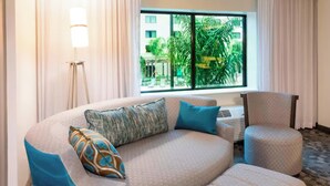 A hotel room with a curved sofa, a chair, a standing lamp, and a large window with a view of palm trees and buildings.