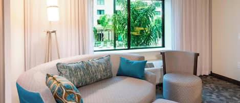 Habitación de hotel con sofá curvo, una silla, una lámpara de pie y un ventanal con vistas a palmeras y edificios.