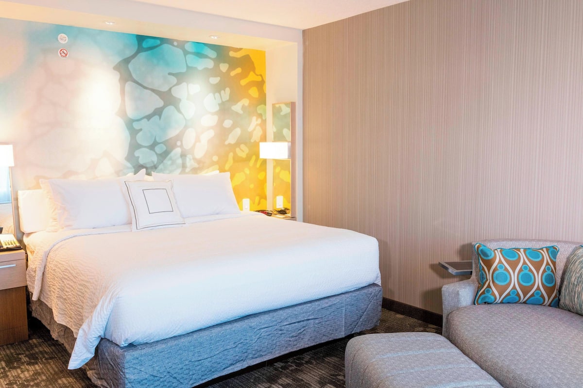 Een hotelkamer met een groot bed, een nachtkastje, een lamp, een stoel en een muur met een kleurrijk abstract ontwerp.