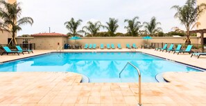 Una piscina rectangular con solárium alicatado, rodeada de sillas reclinables azules y palmeras.
