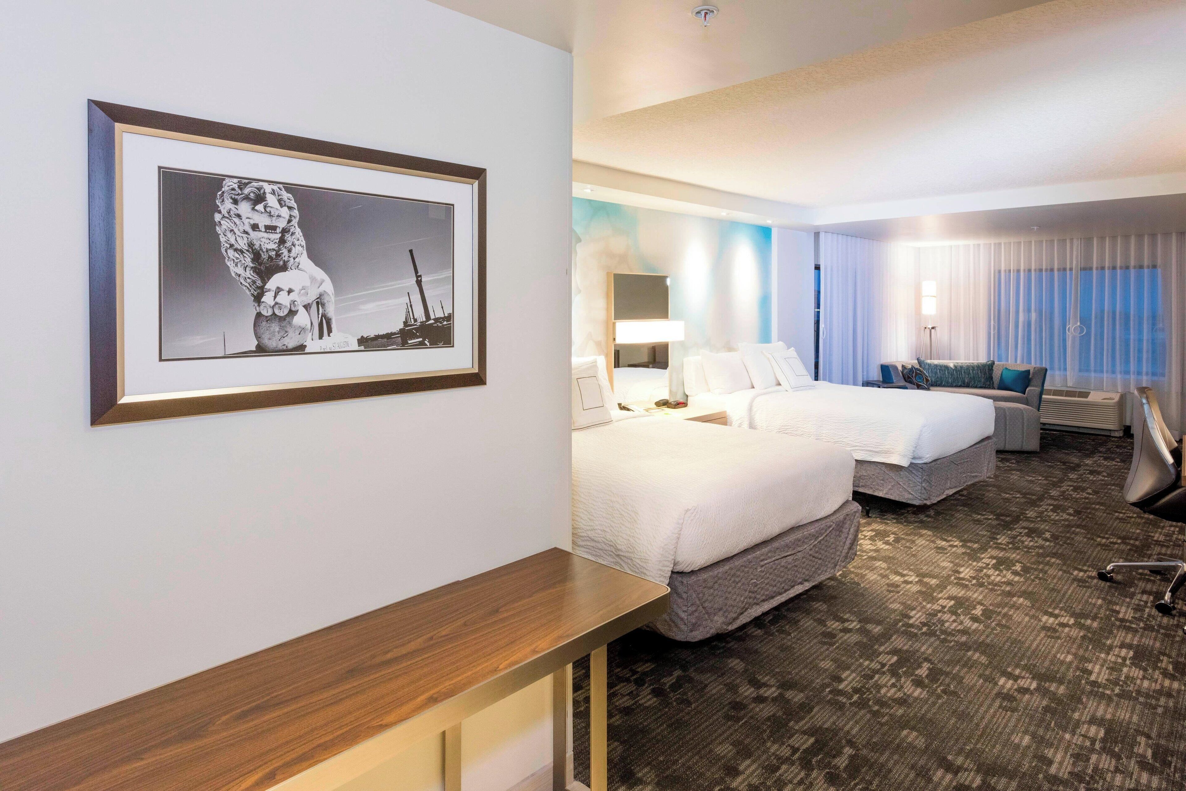 A hotel room with two beds, a desk, a chair, a framed picture on the wall, and a large window with curtains.