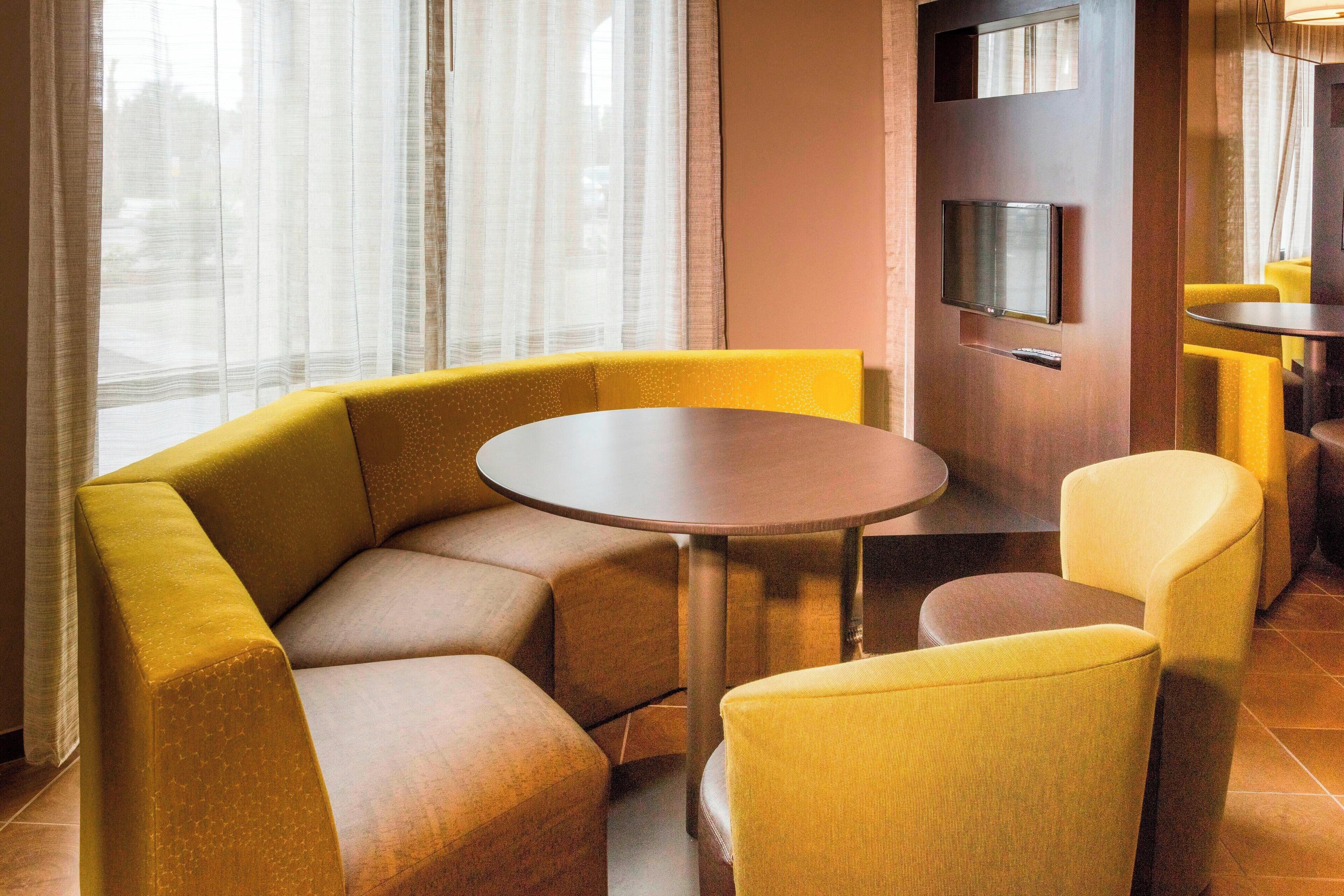 A corner seating area with a round table and yellow chairs, a flat-screen TV mounted on a wall, and a window with sheer curtains.