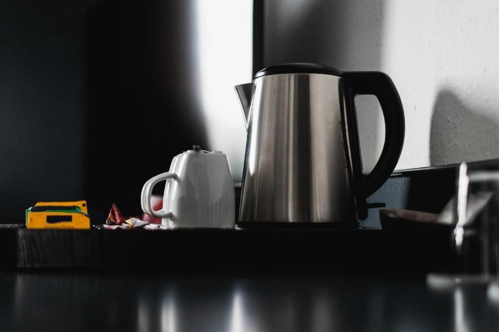 Una cafetera de acero inoxidable y una taza blanca sobre una superficie oscura, con un objeto amarillo y algunos papeles cerca.