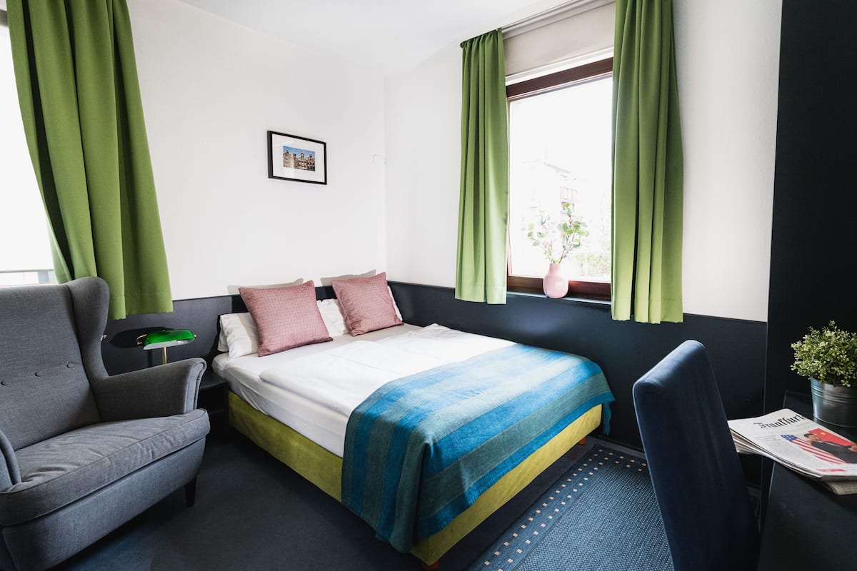 A hotel room with a bed, a grey armchair, a small table with a newspaper, and a window with green curtains.