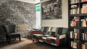 A room with a stone wall, a black armchair, a multicolored sofa, a small table with a lamp, and a bookshelf filled with books. - City Hotel Fellbach (Fellbach)