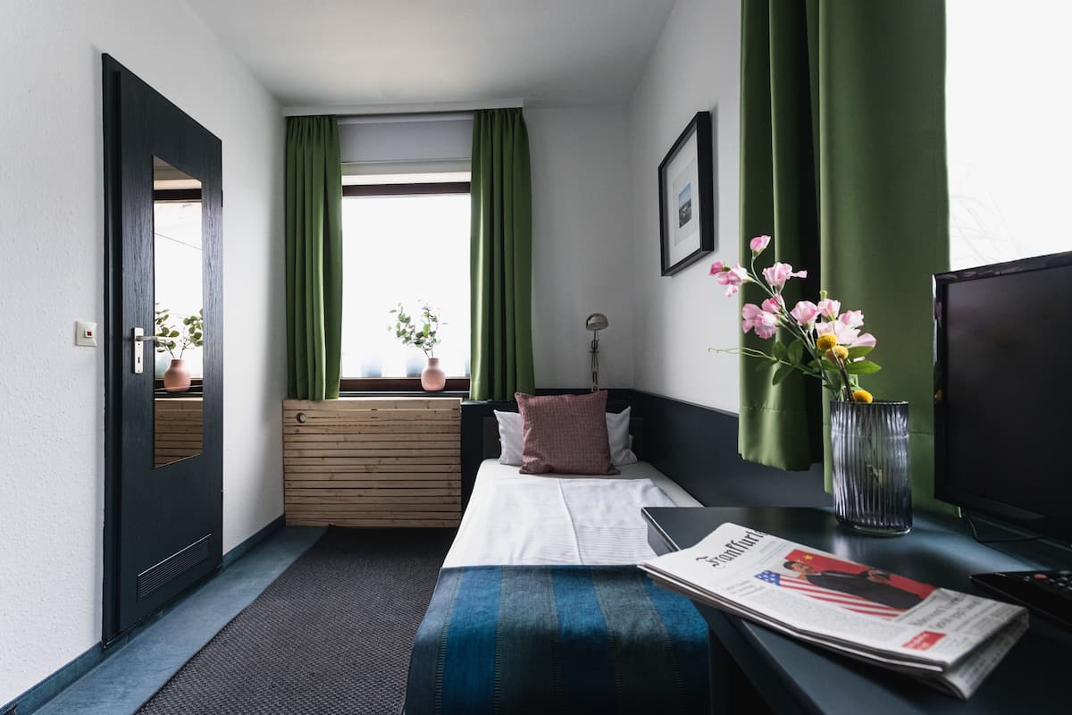 A hotel room with a bed, a desk with a computer, a window with green curtains, a vase with flowers, and a framed picture on the wall.