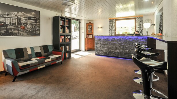 A modern living room with a sofa, a bookshelf, a wooden cabinet, and a bar area with stools. - City Hotel Fellbach (Fellbach)
