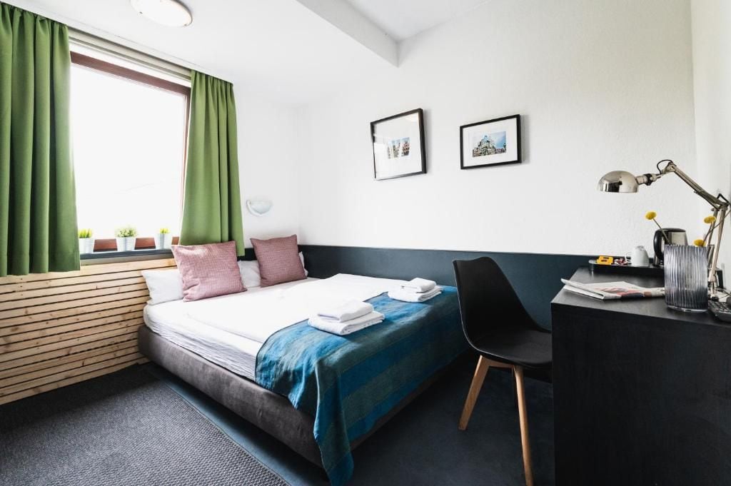 Une chambre d’hôtel comprenant un lit, un bureau, une chaise, une fenêtre avec des rideaux et deux tableaux encadrés accrochés au mur.