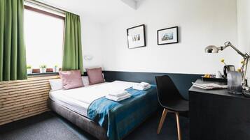 Una habitación de hotel con cama, escritorio, silla, ventana con cortinas y dos cuadros en la pared.