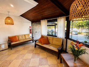 A reception area with a wooden ceiling, two sofas with cushions, a fire extinguisher, a window with curtains, and a vase with flowers. - Búzios Centro Hotel (Búzios)
