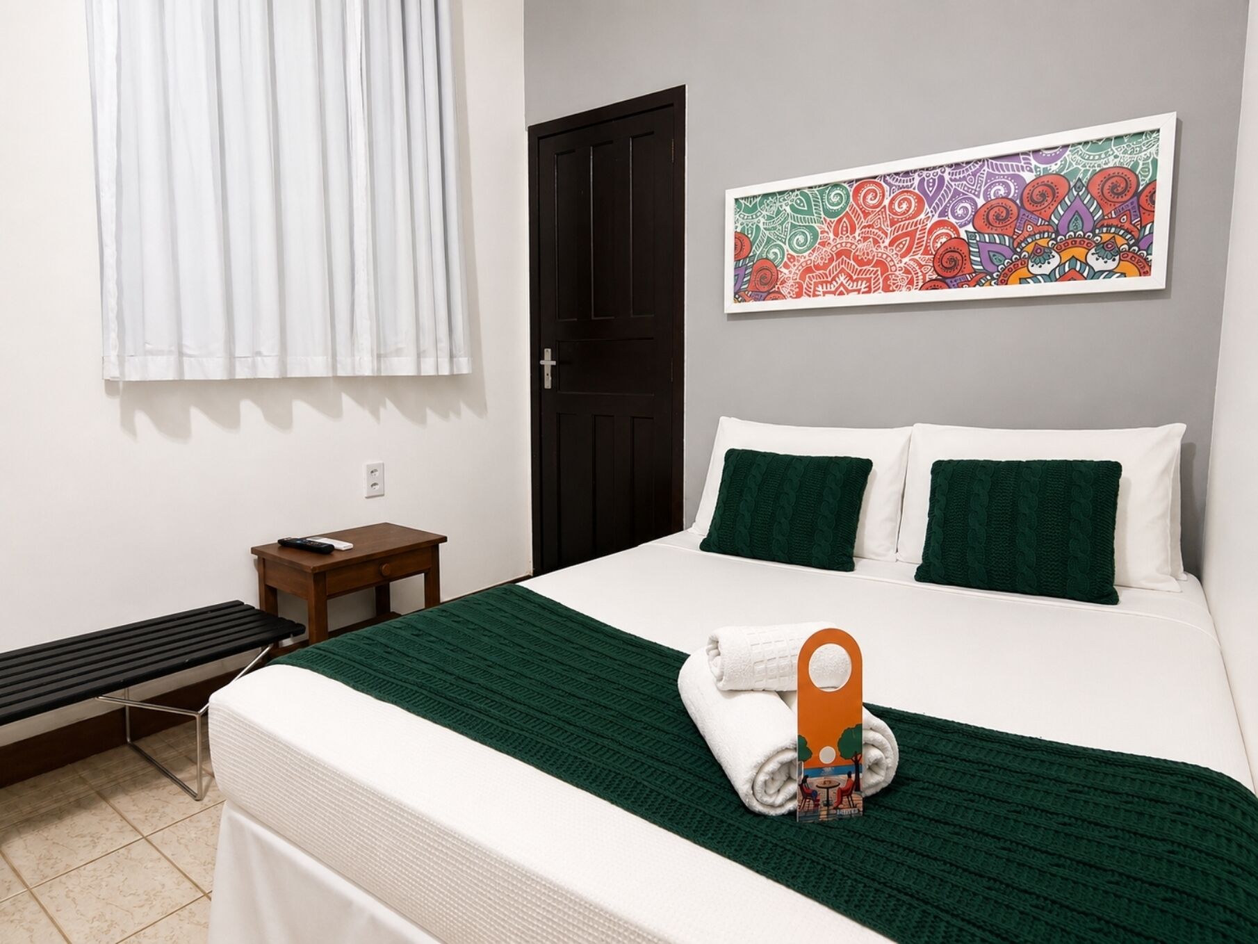 A hotel room with a bed, a small bedside table, a bench, a door, and a colorful artwork on the wall.