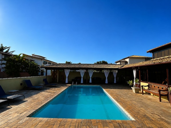 Piscina rectangular con solárium empedrado, rodeada de sillas de descanso y una zona de estar cubierta con cortinas blancas.