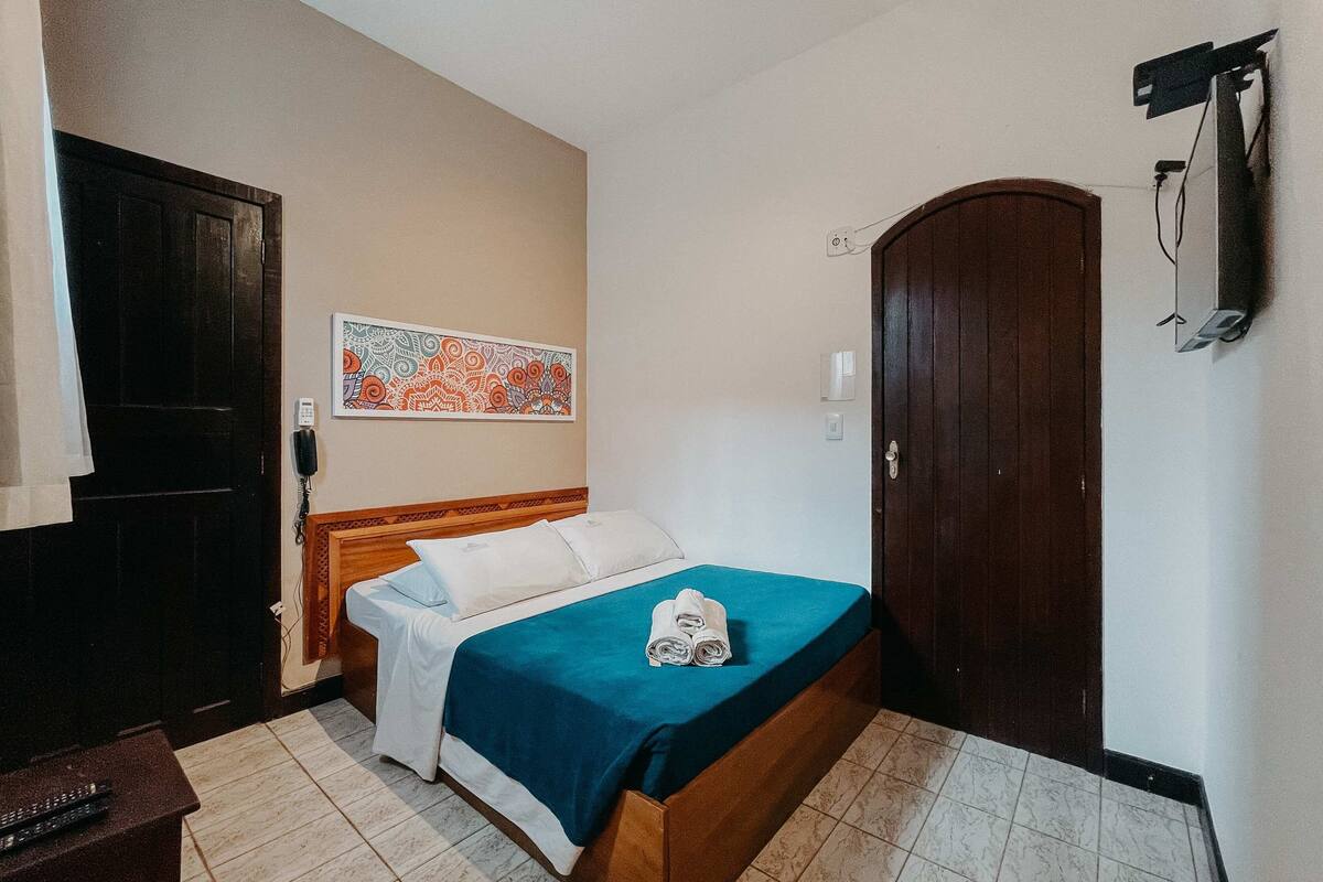 A hotel room with a bed, a wooden headboard, a blue bedspread, a white pillow, a wall-mounted TV, and a door.