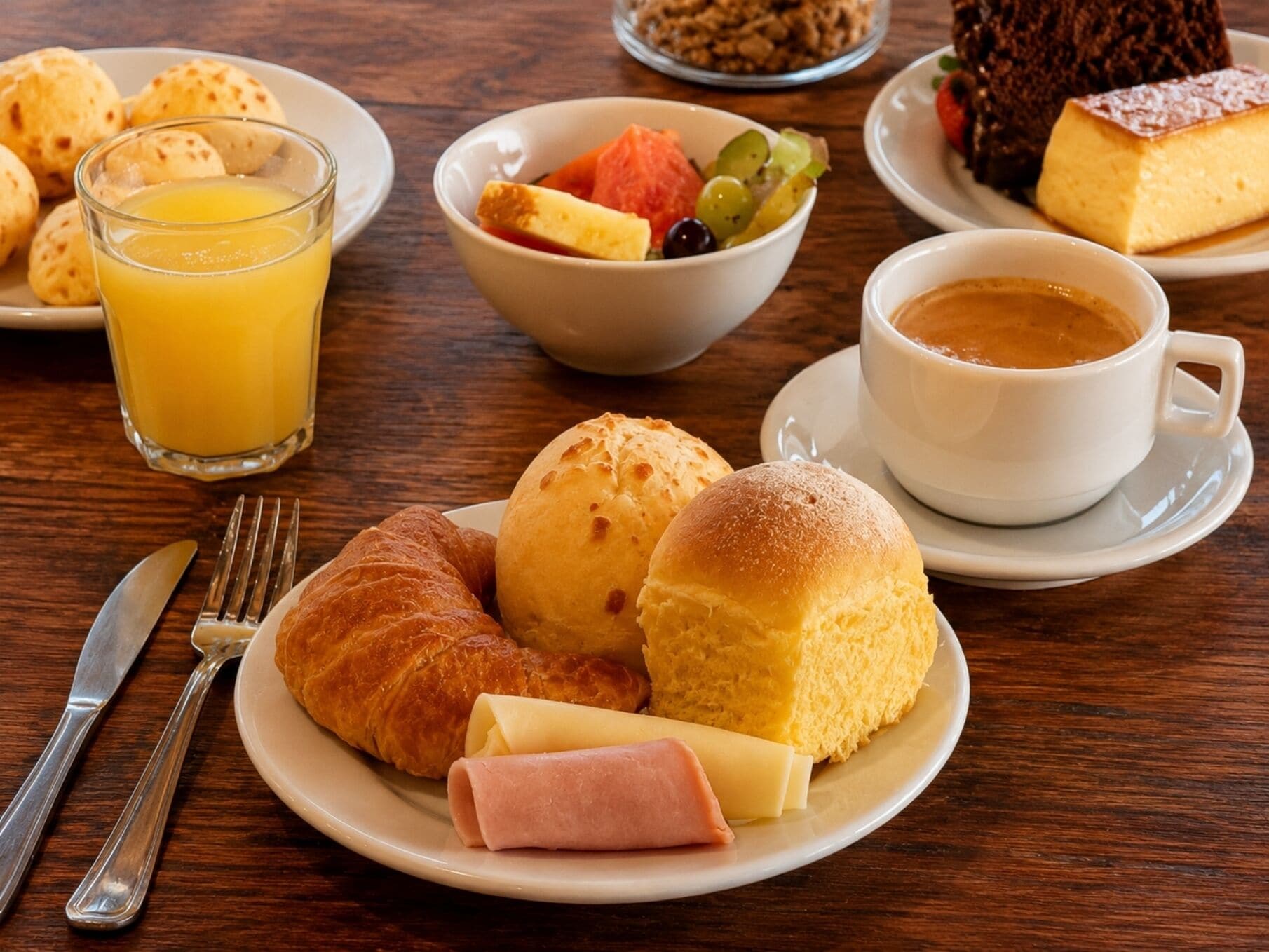 A breakfast setting with croissants, ham, cheese, fruit, and a cup of coffee on a wooden table.