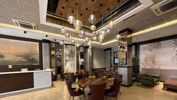 Un hall d’hôtel moderne avec un comptoir d’accueil en bois, une table de salle à manger ronde entourée de chaises en cuir, un canapé vert et une grande fresque murale.