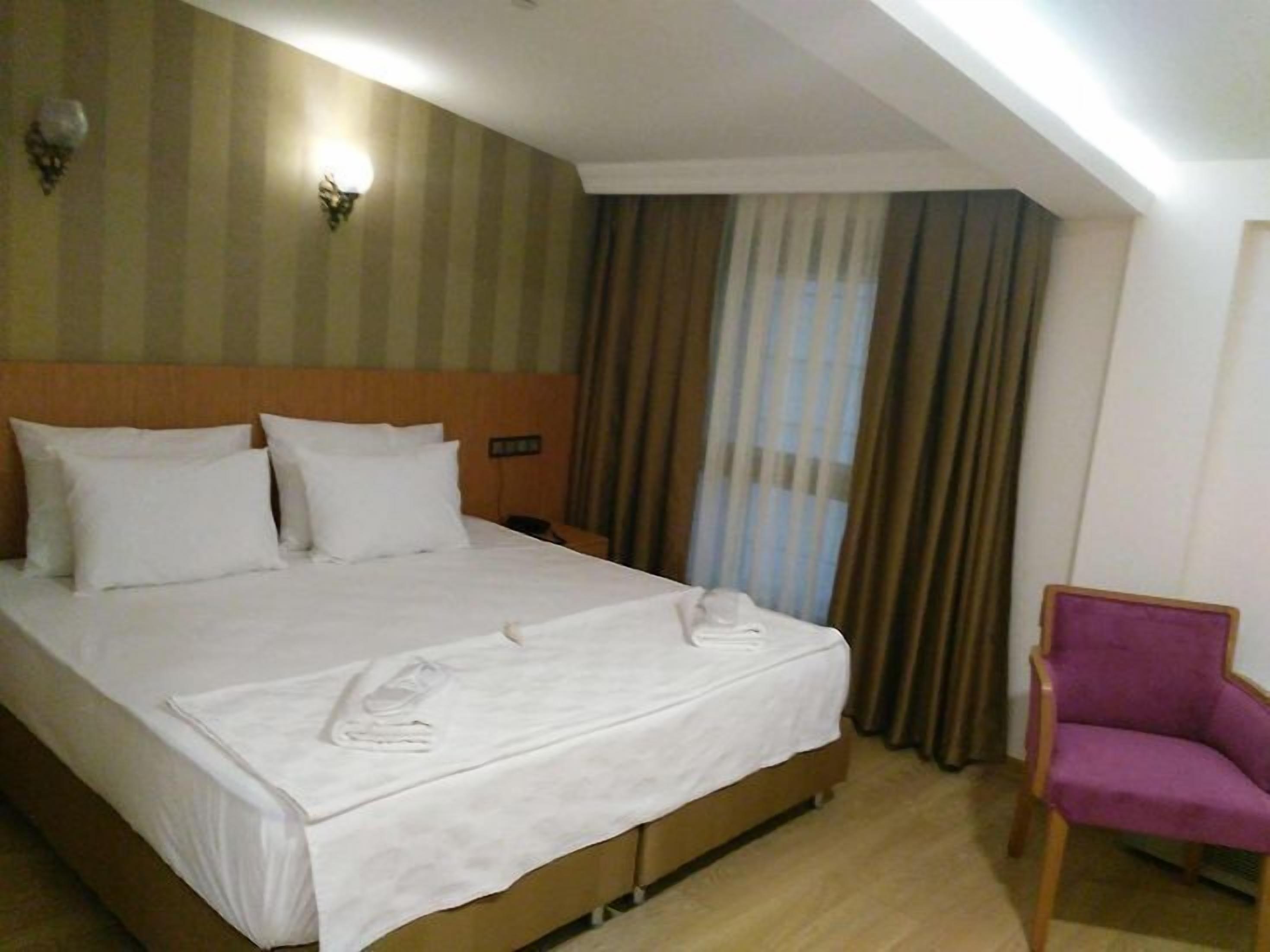 A hotel room with a large bed, a wooden headboard, a nightstand, a purple chair, and a window with curtains.