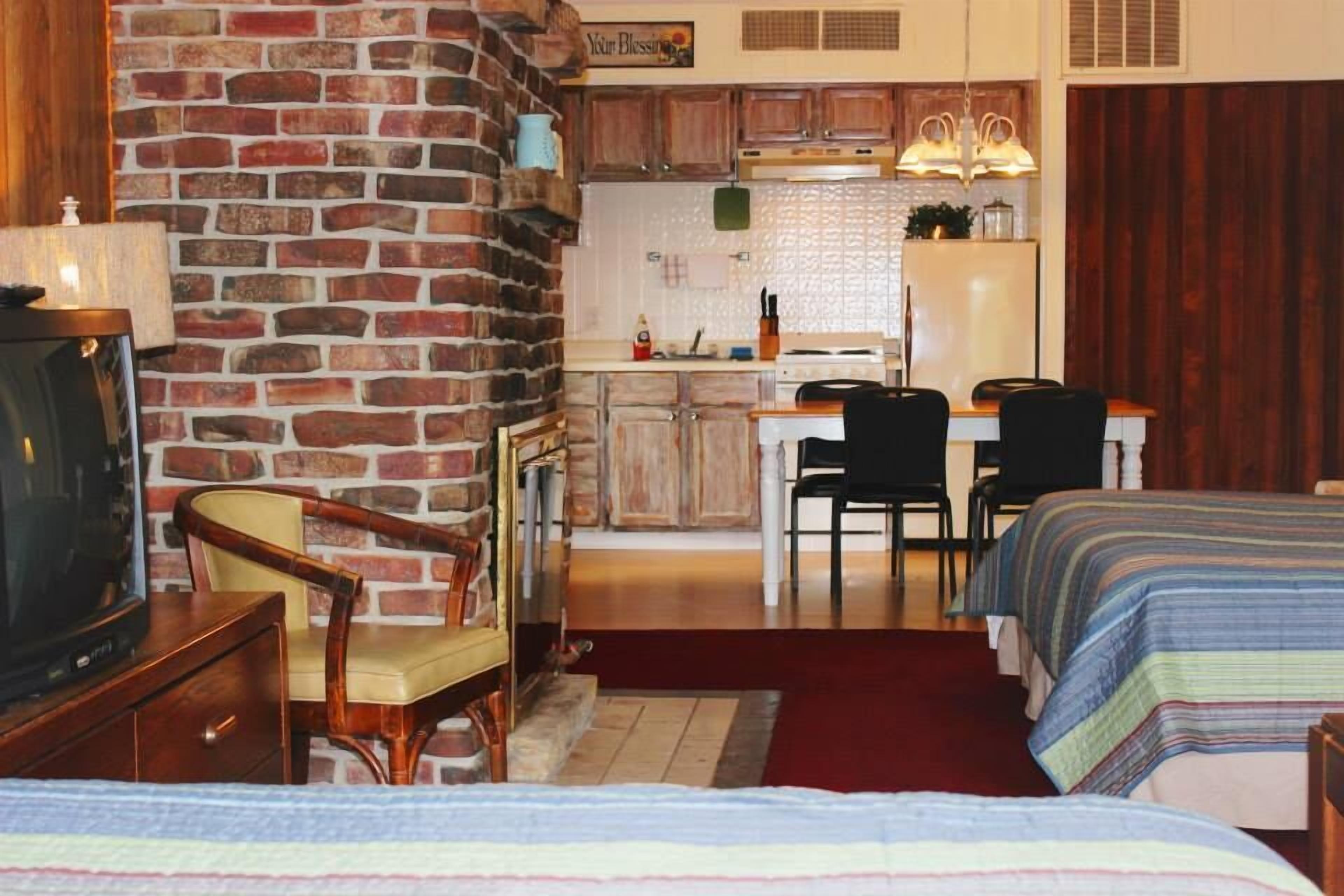 Een gezellige kamer met een bakstenen muur, een televisie, twee bedden met streepjesbeddengoed, een houten stoel, een kleine tafel en een kitchenette met een koelkast en kastjes.
