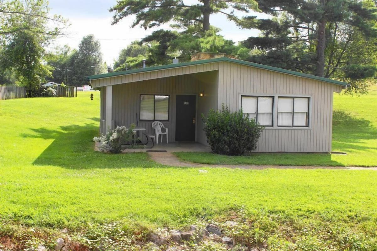 A single-story, light-colored building with a green roof, a front door, and a small porch with two chairs.