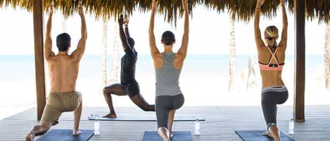Three people doing yoga on mats under a thatched roof, with a beach in the background.