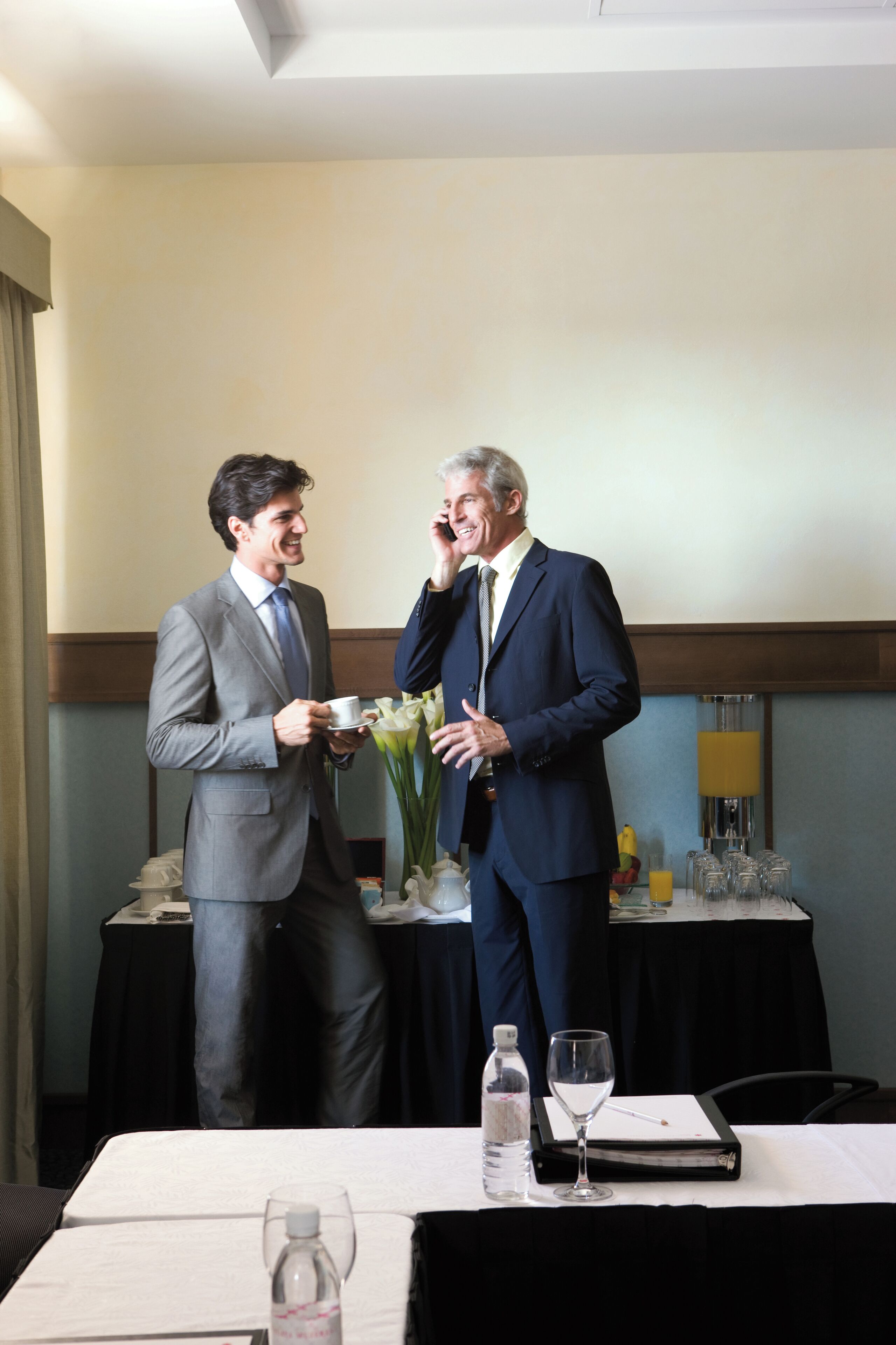 Two men in suits conversing in a hotel room with a table set for a meal, a vase with flowers, and a bottle of water.