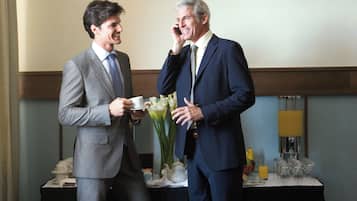 Two men in suits conversing in a hotel room with a table set for a meal, a vase with flowers, and a bottle of water.