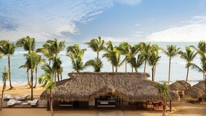 A beach resort with thatched-roof huts, palm trees, and a sandy beach. - Excellence Carmen Punta Cana - Adults Only All Inclusive (Punta Cana)