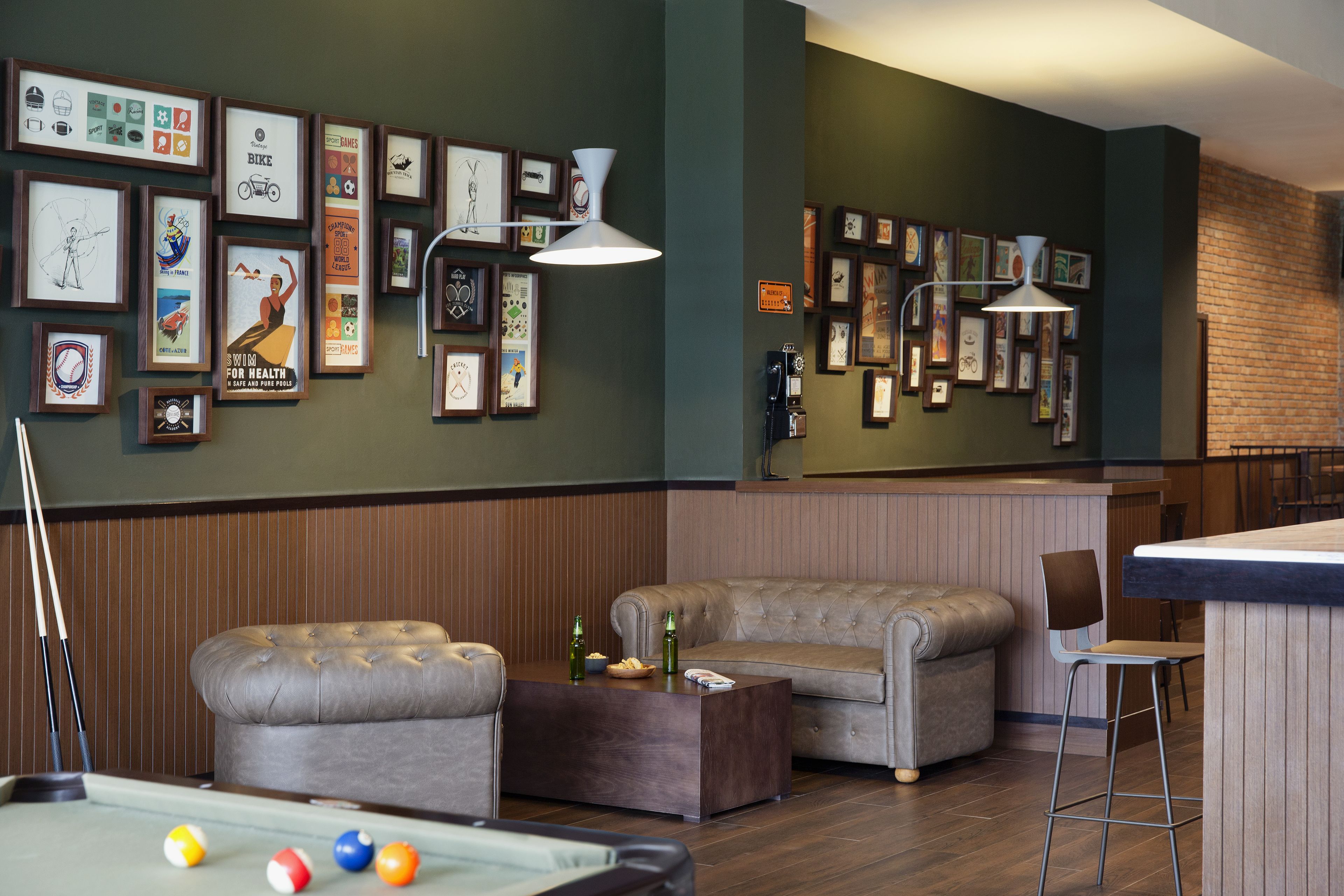 A billiards table with balls, two cushioned sofas, a bar area with stools, and a wall adorned with framed pictures.