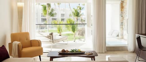 A modern living room with a sofa, a coffee table with fruits, and a view of a pool and palm trees.