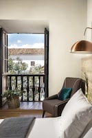 Un balcón con vistas a edificios y árboles, una silla marrón con un cojín azul verdoso, un sofá blanco y una lámpara colgante de cobre.