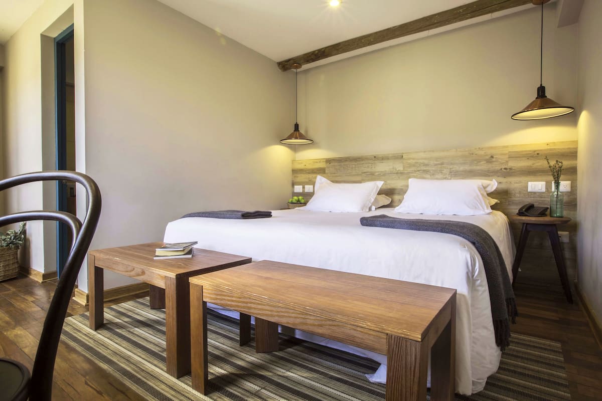 Un dormitorio ordenado con cabecera de madera, cama con ropa de cama blanca, mesita de noche con lámpara y una silla.