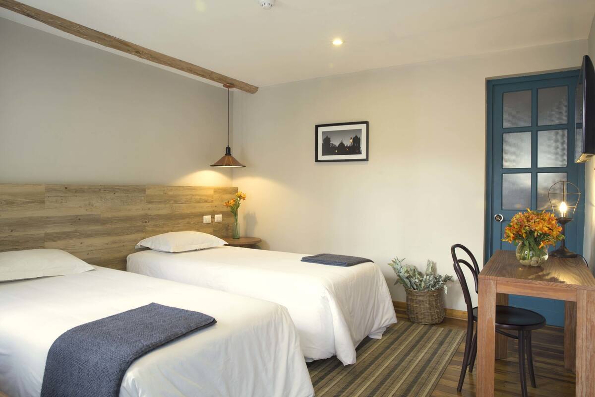 Habitación de hotel con dos camas individuales, cabecera de madera, una mesita con un florero, una silla e imagen enmarcada en la pared.