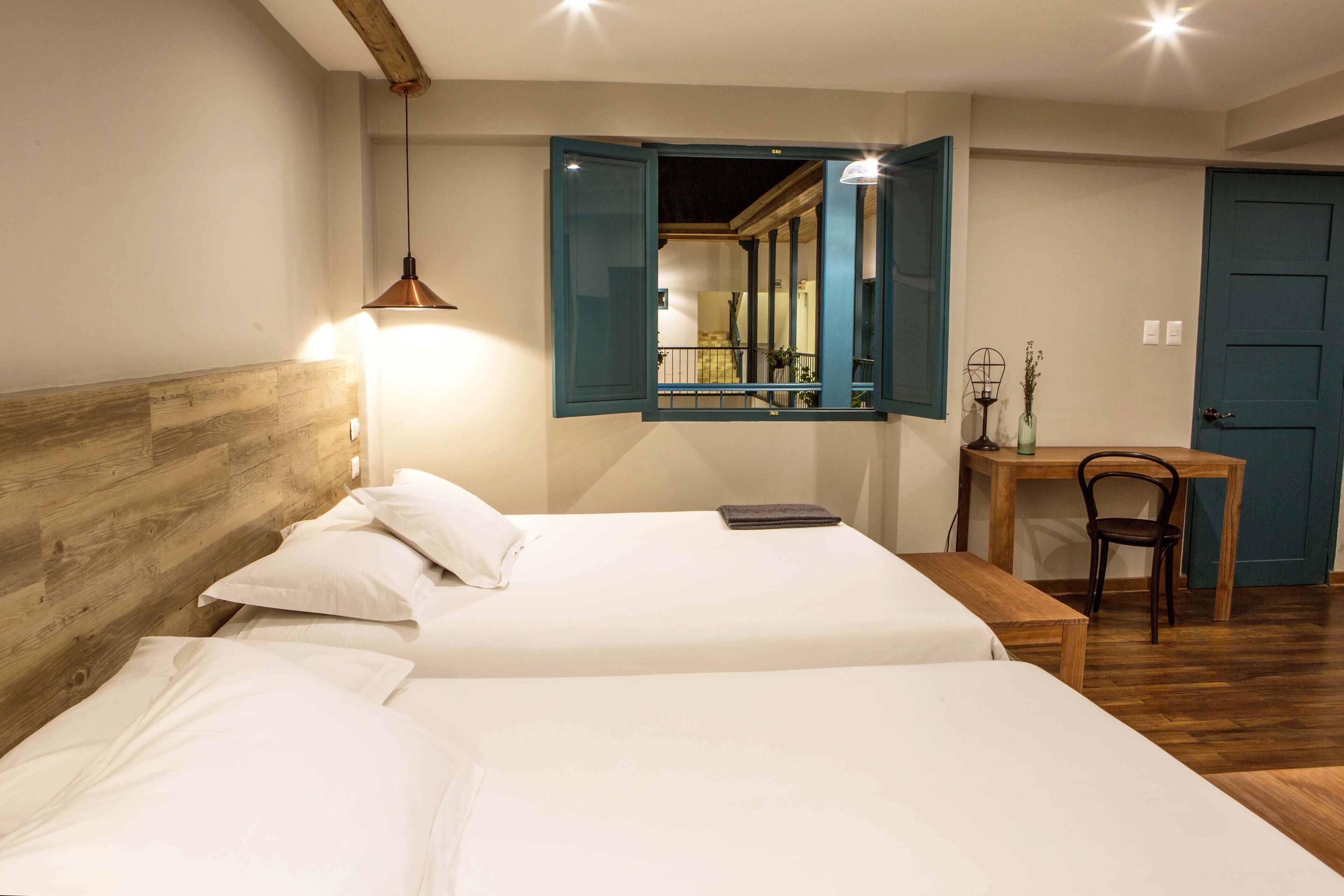 Habitación de hotel con dos camas, cabecera de madera, escritorio con silla y espejo con persiana abierta.