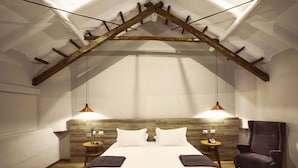 A bedroom with a sloped ceiling, wooden beams, a bed with white linens, two bedside tables with lamps, and a chair. - Abittare Hotels (Cusco)