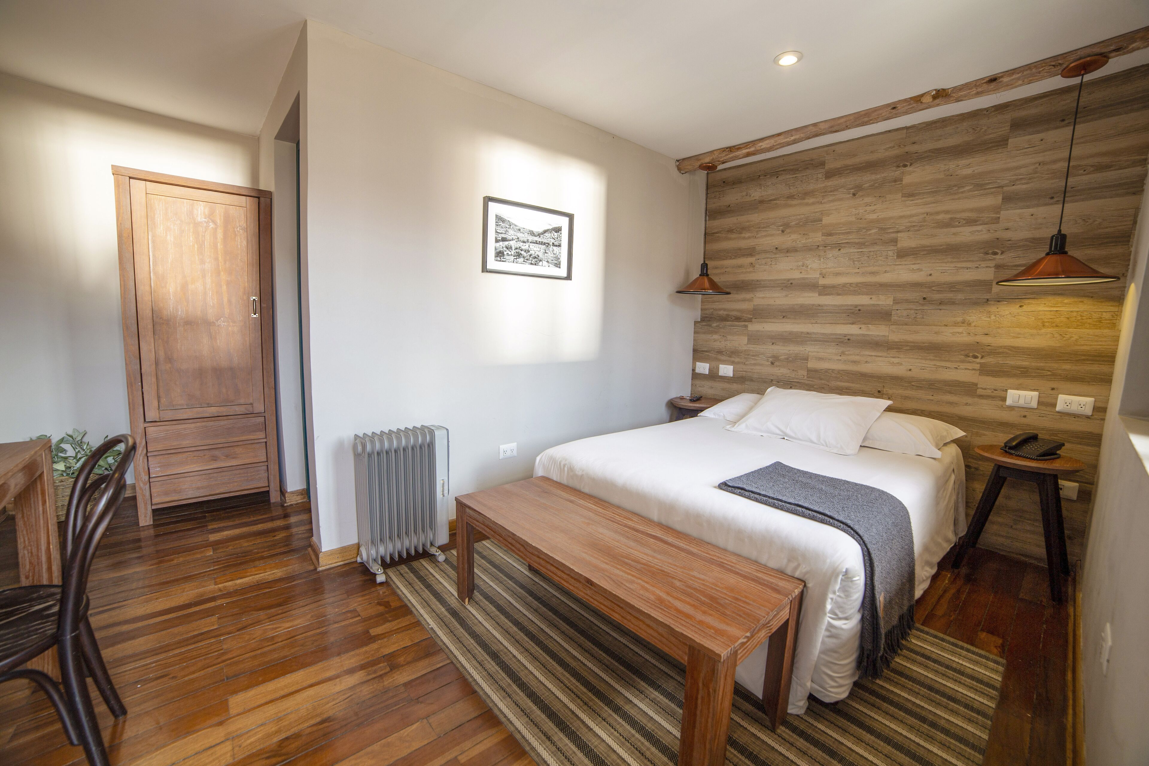 A bedroom with a wooden headboard, a bed with white linens, a wooden bench, a radiator, and a picture on the wall.