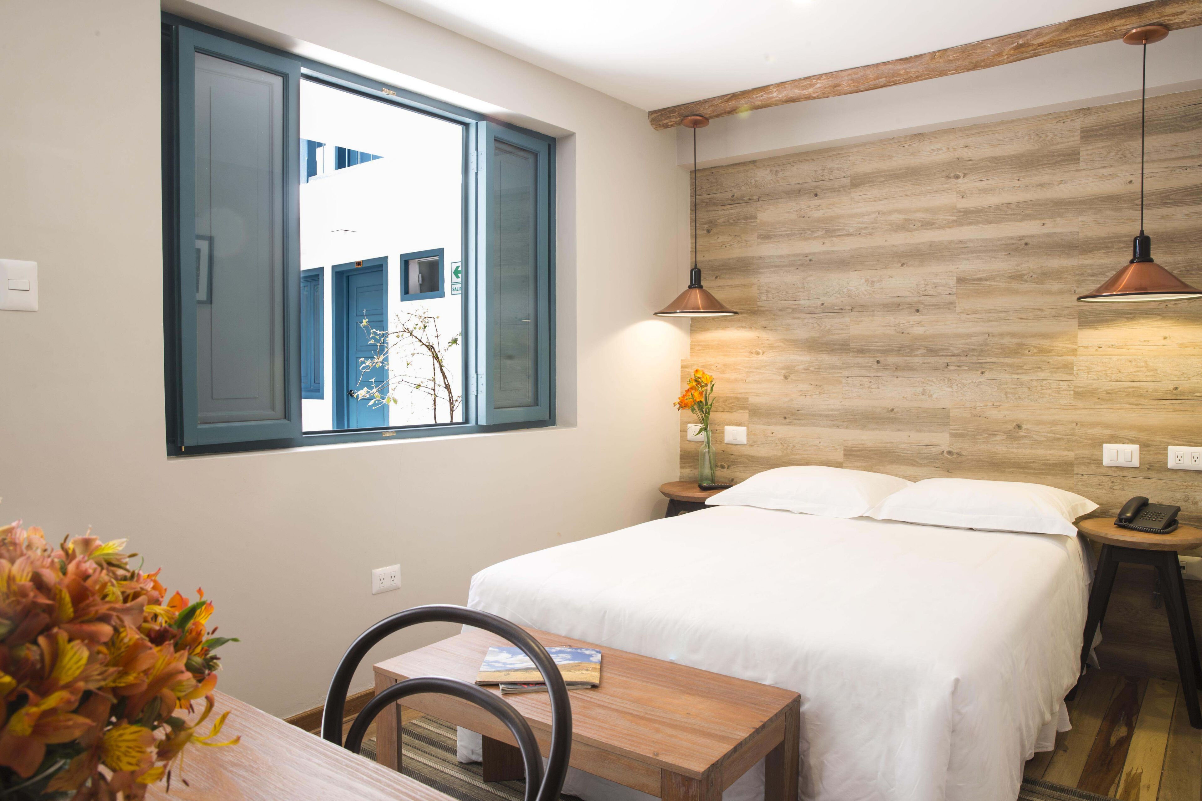 A bedroom with a bed, bedside table, lamp, and a window with a view of a building.