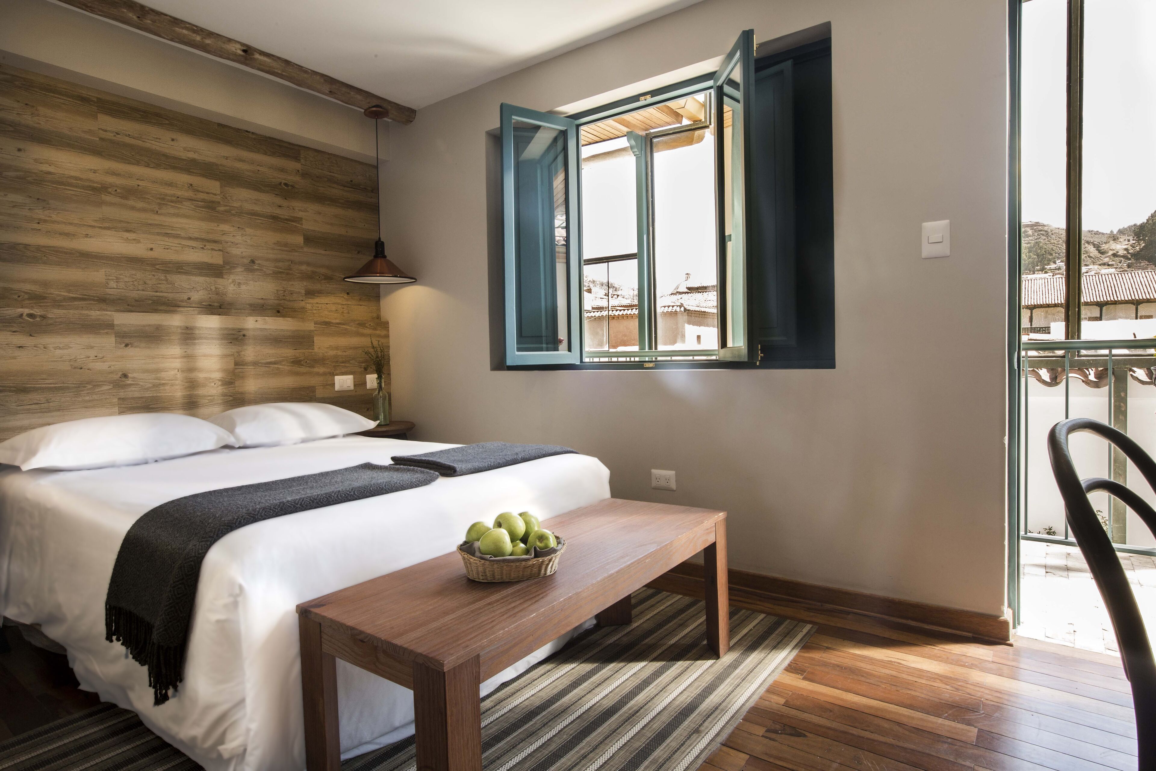 A bedroom with a wooden headboard, a bed with white linens, a wooden nightstand, a wicker basket with apples, a wooden bench, and a window with blue shutters.