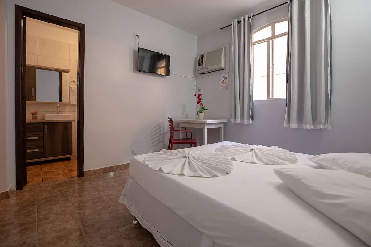 A hotel room with a bed, a desk, a chair, a TV, an air conditioner, and a bathroom with a sink and mirror.