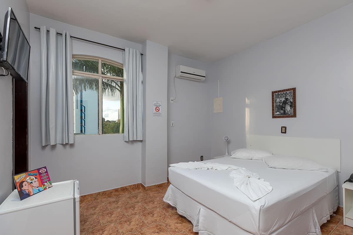 A hotel room with a bed, a TV, a window with curtains, an air conditioning unit, and a framed picture on the wall.