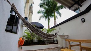 Un hamac tendu entre deux bâtiments, un banc en bois, une terrasse carrelée, un évier blanc et un palmier.