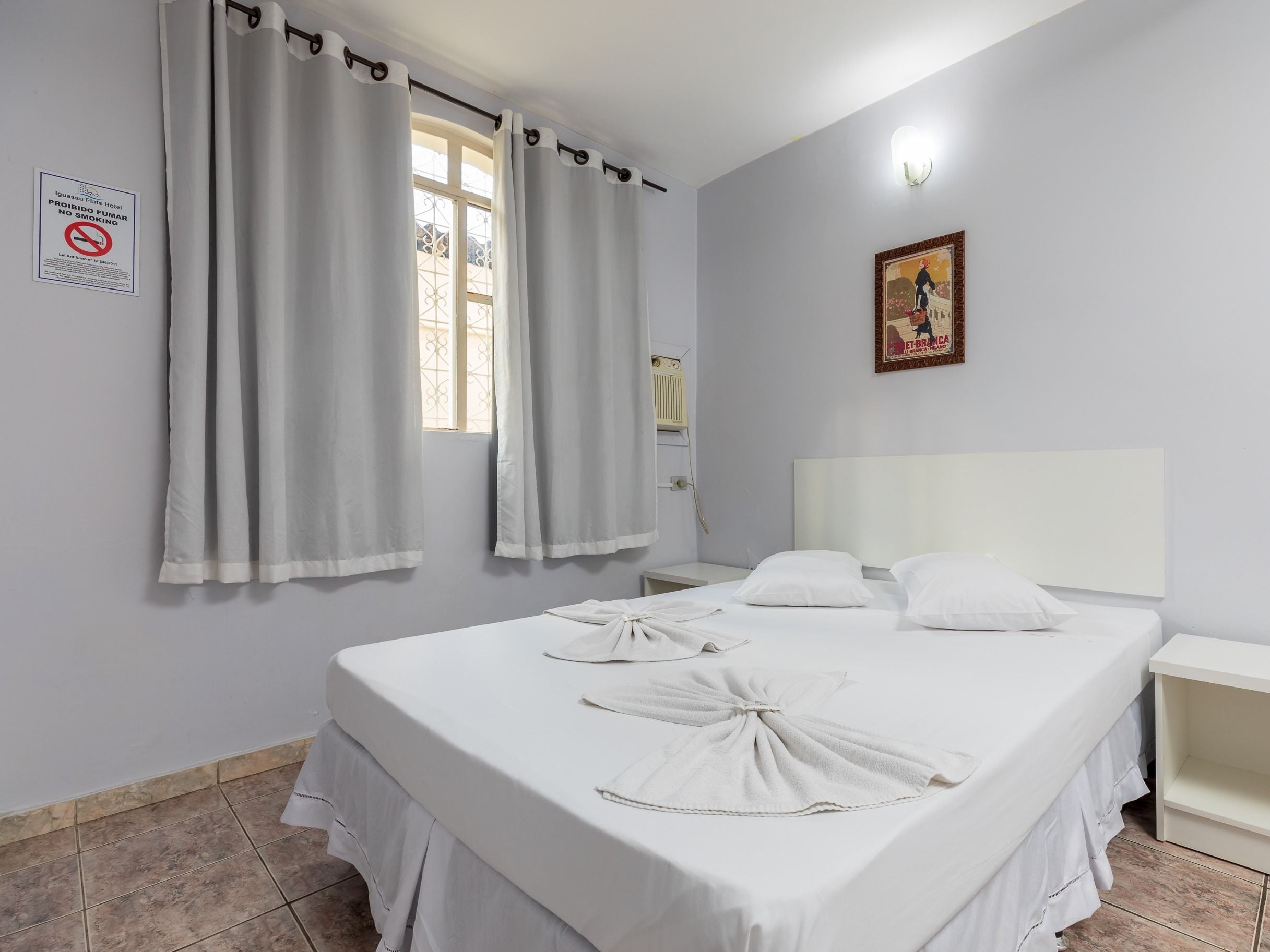 Une chambre d’hôtel avec un lit, des rideaux, une lampe fixée au mur, un tableau et un panneau « défense de fumer ».