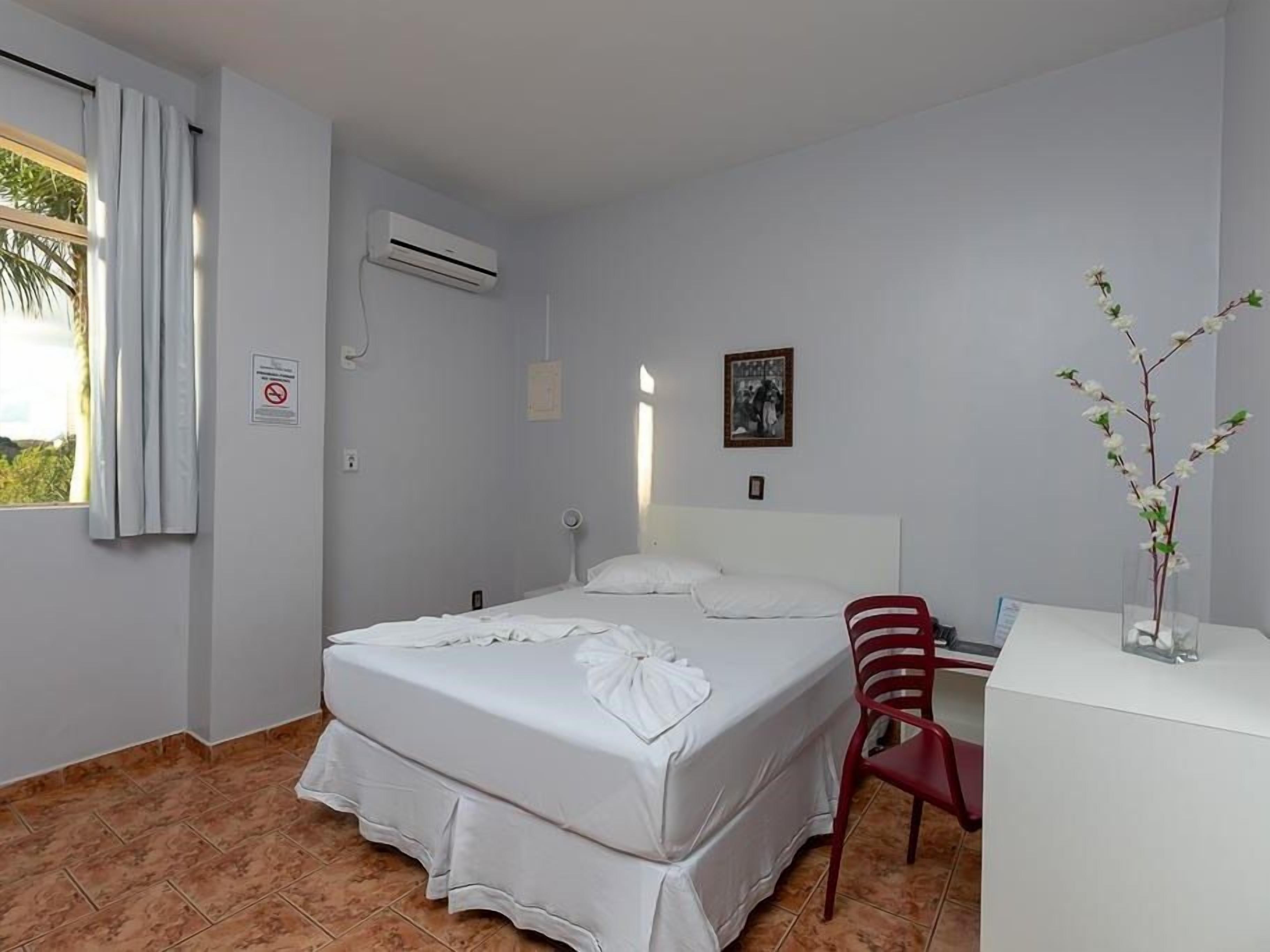Quarto de hotel com cama, escrivaninha, cadeira, ar condicionado e janela com cortinas.