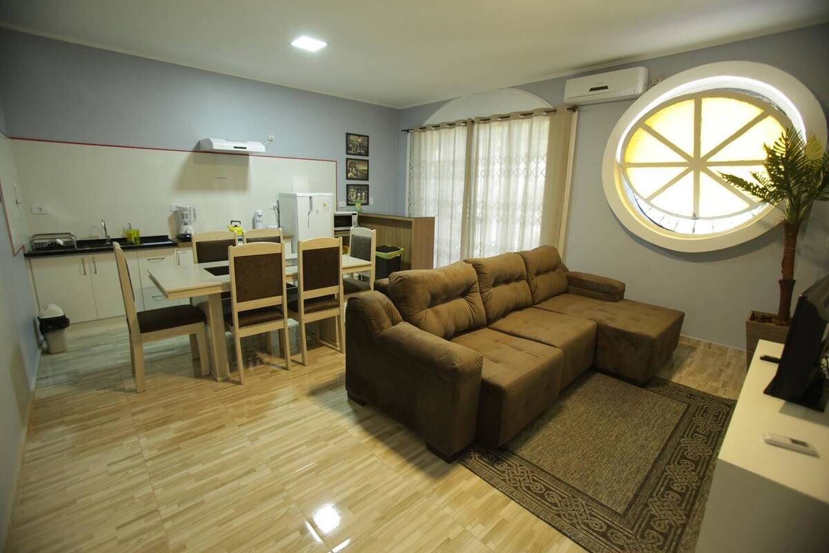 A compact living space with a dining area, a sofa, a round window, and a kitchen in the background.