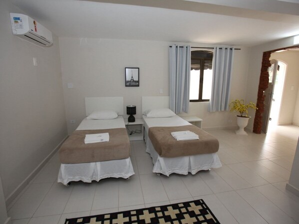 A hotel room with two beds, a bedside table, a lamp, a window with curtains, and a potted plant.