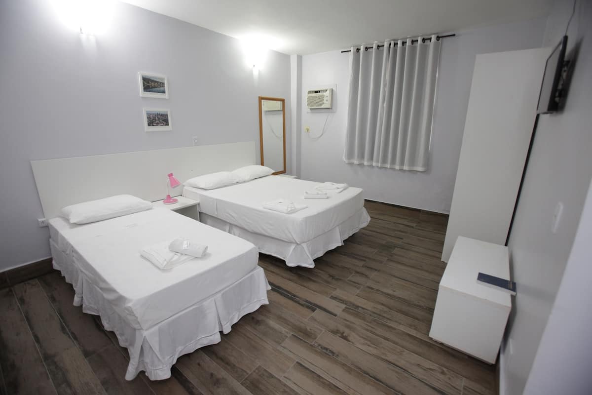 A hotel room with two single beds, a small table, a mirror, an air conditioning unit, and a wardrobe.