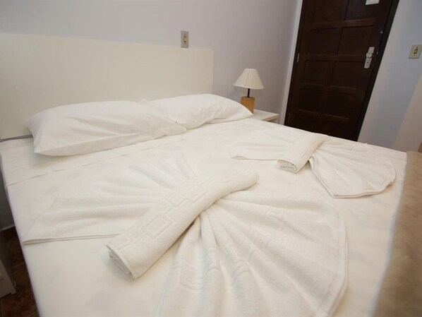 A neatly made bed with a white duvet and pillows, a wooden nightstand with a lamp, and a wooden door in the background. - Iguassu Flats Hotel (Foz do Iguaçu)