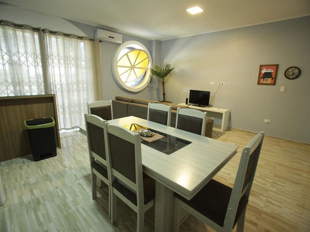 A dining area with a table and chairs, a television on a stand, a window with curtains, and a trash bin.
